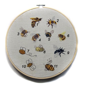 May include: Embroidered hoop art featuring 11 different bees, each numbered 1 through 11. The bees are a variety of colors, including yellow, black, and brown. The hoop is made of wood and has a white fabric background.