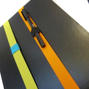 May include: A black notebook with three elastic bands in yellow, orange, and blue. A black pen is tucked into the orange band.