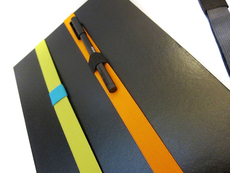 May include: A black notebook with three elastic bands in yellow, orange, and blue. A black pen is tucked into the orange band.