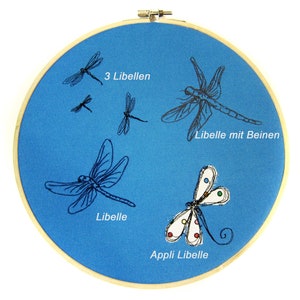 May include: A blue embroidery hoop with four black embroidered dragonflies and one colorful appliqu&eacute; dragonfly. The text "3 Libellen", "Libelle mit Beinen", "Libelle", and "Appli Libelle" is embroidered on the hoop.