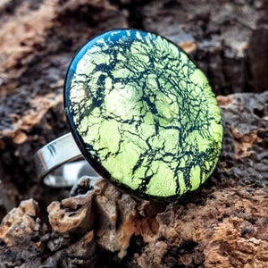 Könnte beinhalten: Silberring mit einem großen, runden Dichroic-Glasstein. Der Stein hat ein limettengrünes und schwarzes Crackle-Muster, das ein einzigartiges und auffälliges Design erzeugt. Der Ring ist vor einem natürlichen, strukturierten Hintergrund platziert.