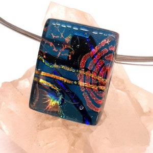 Op de afbeelding: Een rechthoekige hanger gemaakt van dichroïsch glas met een blauw, zwart, rood en gouden patroon. De hanger hangt aan een zilveren ketting.