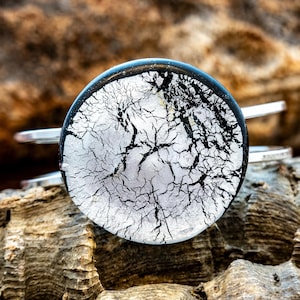 May include: A silver-toned cuff bracelet featuring a round, abstract design. The design has a black and white pattern resembling tree branches or cracks, set against a silver background. The bracelet is displayed on a textured, brown surface.