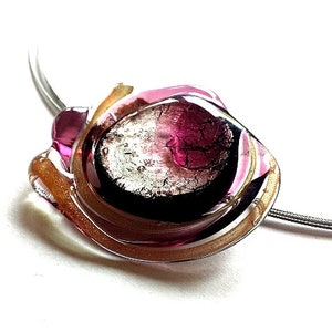 Das Schmuckstück dieser Halskette hat einen Durchmesser von ca. 3-4 cm und wiegt ca. 15g. Der Anhänger ist auf schwarzer Grundfläche mit Farbglas verarbeitet.
Das farbig- transparente Glas ist in den Farben rosa und silber