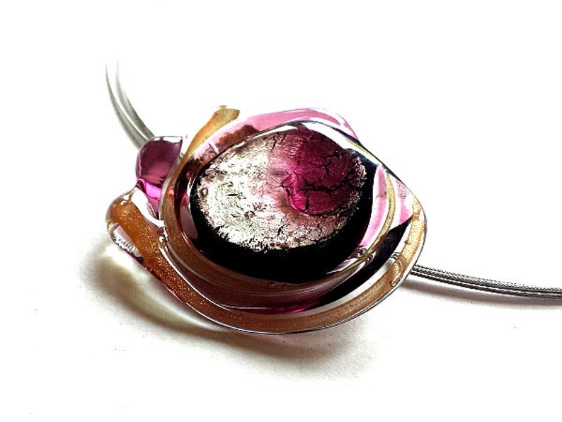 Das Schmuckstück dieser Halskette hat einen Durchmesser von ca. 3-4 cm und wiegt ca. 15g. Der Anhänger ist auf schwarzer Grundfläche mit Farbglas verarbeitet.
Das farbig- transparente Glas ist in den Farben rosa und silber
