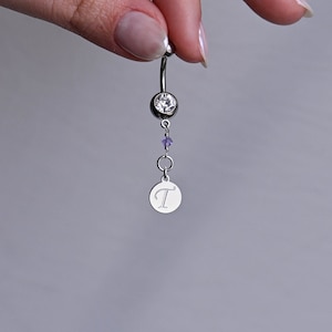 Op de afbeelding: Een zilveren navelpiercing met een heldere kristal, een paarse kristal en een zilveren bedel met de letter 'T' erop gegraveerd.