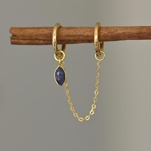 May include: A pair of gold-colored hoop earrings connected by a delicate chain. One earring features a small, dark blue, marquise-shaped gemstone pendant. The earrings are displayed on a brown, cylindrical object against a neutral background.