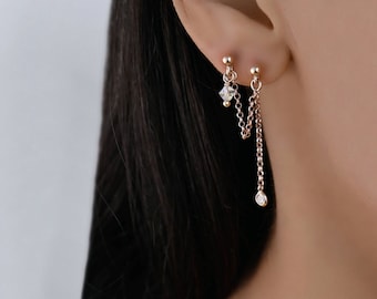 Op de afbeelding: Een paar gouden kettingoorbellen met een klein kristal en een ronde diamant aan het einde van elke ketting. De oorbellen worden in de oorlel en helix piercing gedragen.
