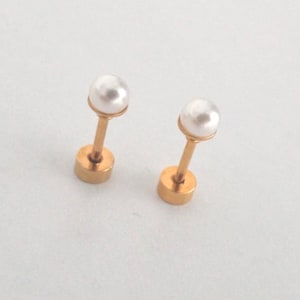 Op de afbeelding: Een paar oorknopjes met ronde witte parels op goudkleurige metalen staafjes. Elke oorbel bestaat uit een gladde, bolvormige parel bevestigd aan een dun, recht metalen steeltje dat uitkomt op een platte, ronde basis aan de onderkant.