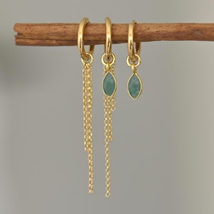Puede incluir: Tres pendientes de aro dorados con diferentes diseños. Un par tiene una larga cadena dorada, y los otros dos tienen una piedra en forma de lágrima, de color verde claro. Los pendientes se muestran en un palo marrón sobre un fondo gris.
