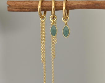 May include: Three gold hoop earrings with different designs. One pair has a long gold chain, and the other two have a teardrop-shaped, light-green stone. The earrings are displayed on a brown stick against a gray background.
