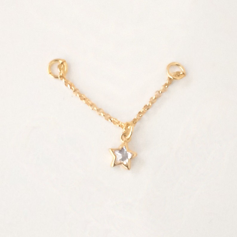 K&ouml;nnte beinhalten: Eine goldene Kette mit zwei Sprungringen und einem Sternanh&auml;nger mit einem klaren Kristall in der Mitte.