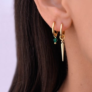 Peut inclure: Boucles d'oreilles créoles en or avec un charme en cristal vert et un charme en pointe dorée suspendu en bas.