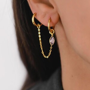 Puede incluir: Pendientes de aro dorados conectados por una delicada cadena. Un pendiente presenta una pequeña gema púrpura de forma ovalada. Los pendientes están diseñados para usarse en perforaciones adyacentes en las orejas, creando un aspecto elegante y conectado.