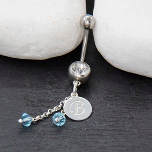 Op de afbeelding: Een zilveren navelpiercing met een heldere edelsteen en een bungelende bedel. De bedel bevat een kleine schijf met de letter "B" en twee lichtblauwe kristallen kralen. De sieraden worden tegen een donkere achtergrond getoond.