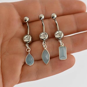 Op de afbeelding: Drie zilverkleurige, gebogen navelpiercings, elk voorzien van een heldere ronde kristal aan de bovenkant en een bungelende lichtblauwe steen. De stenen hebben de vorm van een druppel, een marquise en een rechthoek, verbonden door metalen ringetjes.