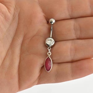 May include: A silver belly button ring with a clear, round gem and a dangling, oval-shaped red gem. The top ball and the gem settings are silver. The red gem is approximately 0.75 inches long.