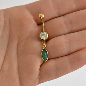 Può includere: Anello per ombelico dorato con un cristallo trasparente e un pendente di pietra preziosa verde.