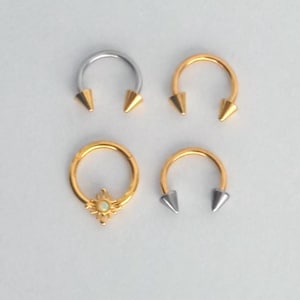 May include: A collection of four body jewelry pieces including three horseshoe-shaped rings with pointed ends and one circular hoop ring. The set features two gold-toned horseshoe rings, one silver-toned horseshoe ring, and one gold-toned hoop ring with a central sunburst design containing a small white stone.