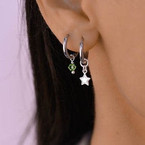May include: A pair of silver hoop earrings with a green crystal charm on one and a silver star charm on the other.