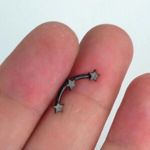 Può includere: Un gioiello per il corpo a forma di bilanciere curvo nero con tre piccoli accenti a forma di stella lungo l'arco. Il metallo è nero opaco e il design consiste in una barra curva liscia con stelle fissate a intervalli lungo il bordo esterno.