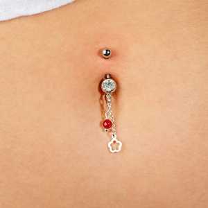 May include: A silver belly button ring with a dangling chain. The top ball is smooth, while the bottom piece features a clear gem, two red beads, and a flower charm. The jewelry is set against a skin tone background.