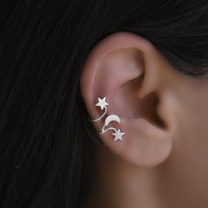 Könnte beinhalten: Silberner Ohrring mit einem Halbmond und zwei Sternen. Der Ohrring ist so konzipiert, dass er sich um den oberen Teil des Ohrs legt.