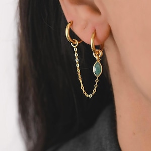May include: A pair of gold-coloured double hoop earrings linked by a fine chain. One earring has a teardrop-shaped, emerald-green gemstone. The earrings are designed to be worn in adjacent ear piercings, creating a stylish, connected appearance.