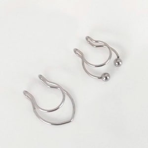 Könnte beinhalten: Zwei silberfarbene Nasenmanschetten aus Metall. Eine Manschette hat ein einfaches Doppelschleifen-Design, während die andere eine Doppelschleifen-Struktur mit zwei kleinen, abgerundeten Kugelenden aufweist. Beide Teile bestehen aus dünnem, poliertem Draht.