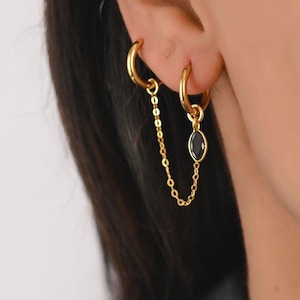 Op de afbeelding: Goudkleurige dubbele oorringen. Eén ring is met een fijne ketting verbonden met een tweede ring. De tweede ring heeft een zwarte, ovaalvormige edelsteen hanger. De oorbellen worden op een oor gedragen.