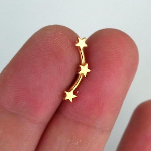 Può includere: Un orecchino a barra curva color oro con tre piccole stelle a cinque punte disposte in fila verticale lungo l'asta metallica. Il gioiello presenta una superficie lucida e riflettente con un design leggermente arcuato.