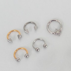 May include: A collection of five body jewelry pieces including two gold-tone circular barbells with ball ends, one silver-tone circular barbell with spike ends, one silver-tone circular barbell with ball ends, and one silver-tone hinged hoop ring with a sun charm.