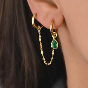 May include: Gold-colored hoop earrings connected by a delicate chain. One earring features a teardrop-shaped, emerald-green gemstone. The earrings are worn on a person's ear, showcasing the jewelry's design and color.
