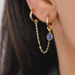 May include: Gold hoop earrings connected by a delicate chain. One earring features a teardrop-shaped, dark blue gemstone. The earrings are designed to be worn in adjacent ear piercings, creating a layered effect. The jewelry is set against a blurred background.