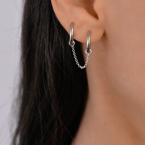 Op de afbeelding: Een paar zilveren oorringen verbonden door een fijne ketting. De oorbellen worden op het oor gedragen, met één ring in de bovenste en de andere in de onderste oor. De ketting hangt tussen de twee ringen.