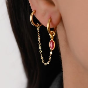 Peut inclure: Boucles d'oreilles créoles dorées reliées par une chaîne. L'une des boucles d'oreilles est ornée d'un pendentif en pierre rouge. L'image met en valeur le design et la couleur des bijoux.