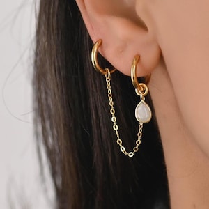 Op de afbeelding: Goudkleurige dubbele oorringen. Eén ring is verbonden met een ketting die naar een tweede ring hangt. De tweede ring heeft een druppelvormige, gebroken witte stenen hanger. De oorbellen worden op het oor gedragen.