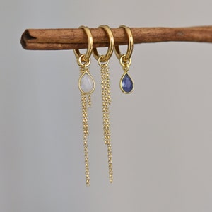 May include: Three gold hoop earrings with dangling chains and gemstone drops. The earrings feature a white moonstone, a blue sapphire, and a clear gemstone.