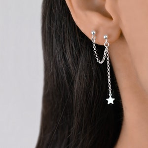 May include: Silver double chain earrings with a star charm hanging from the bottom. The earrings are worn in the upper earlobe.