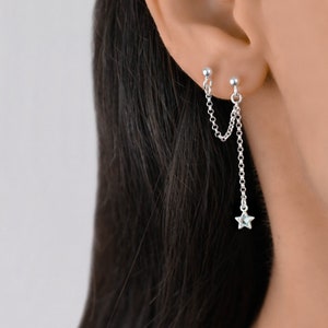 May include: Silver double chain earrings with a star charm hanging from the bottom. The earrings are worn in the upper earlobe.