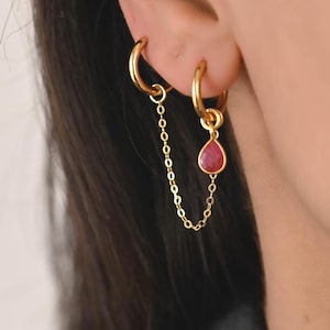 May include: Gold hoop earrings with a chain connecting to a second hoop with a teardrop-shaped, red gemstone. The earrings are worn on a person's ear.