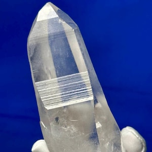 Puede incluir: Un gran cristal de cuarzo transparente con doble terminación. El cristal tiene una superficie natural estriada y se sostiene con un guante blanco.