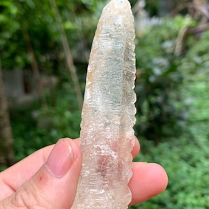 Puede incluir: Un cristal claro y alargado con una superficie rugosa y texturizada. El cristal se sostiene en una mano contra un fondo verde borroso. El color del cristal es un blanco p&aacute;lido y transl&uacute;cido con toques de amarillo y marr&oacute;n.