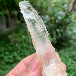 May include: A clear quartz crystal with a rough, textured surface. The crystal is held in a hand, with a blurred green background. The quartz has a natural, irregular shape and a translucent appearance, showcasing its internal structure. The crystal is approximately 10 cm long.