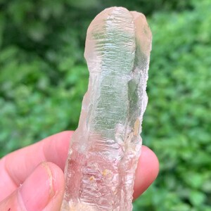 May include: A pale, translucent crystal with a textured surface. The crystal has a light pinkish-beige colour and an irregular shape. The crystal is held in a hand against a blurred green background.