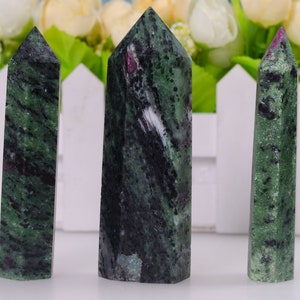 May include: Three green, black, and pink gemstone points, also known as zoisite, are arranged in a row on a white surface. The points are polished and have a smooth, glossy finish.