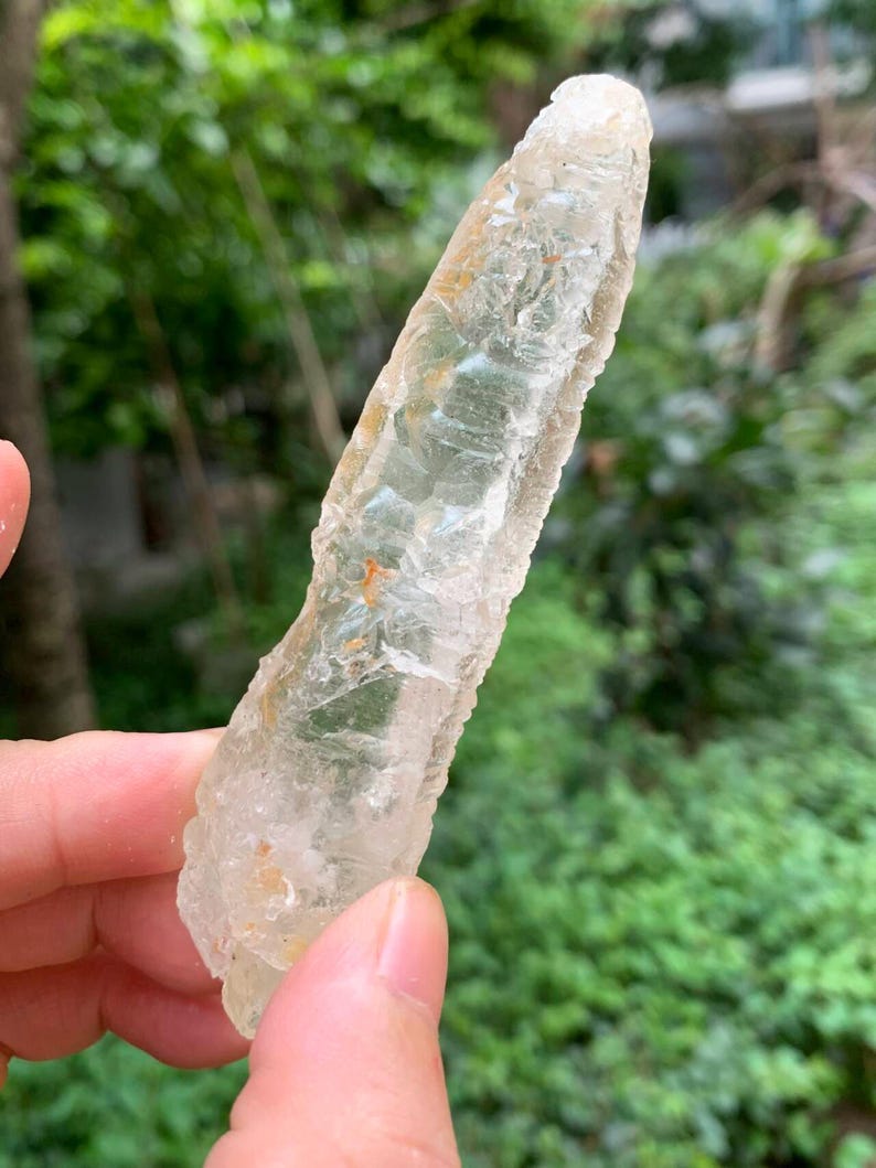 Puede incluir: Un cristal claro y alargado con una superficie rugosa y texturizada. El cristal tiene una apariencia p&aacute;lida y transl&uacute;cida con toques de amarillo y naranja. El cristal se sostiene contra un fondo verde borroso.