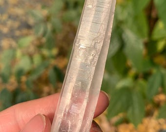 以下が含まれることがあります： 自然なファセット形状の透明な水晶ポイント。ぼやけた緑と茶色の背景に対してクリスタルが保持されています。水晶はわずかにピンクがかった色合いで、内部構造が見えます。