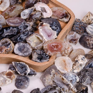 Könnte beinhalten: Eine Sammlung von Achat-Kristallscheiben in verschiedenen Grautönen, Braun- und Weißtönen. Die Kristalle haben eine natürliche, raue Textur und sind in einer Holzschale angeordnet.