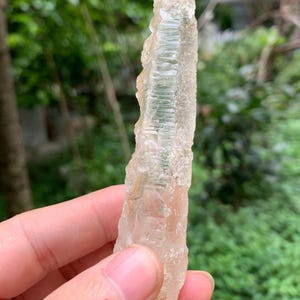 Puede incluir: Una formaci&oacute;n cristalina transl&uacute;cida y clara, con una superficie rugosa y texturizada. El cristal se sostiene en una mano sobre un fondo verde borroso. Es un esp&eacute;cimen mineral natural.